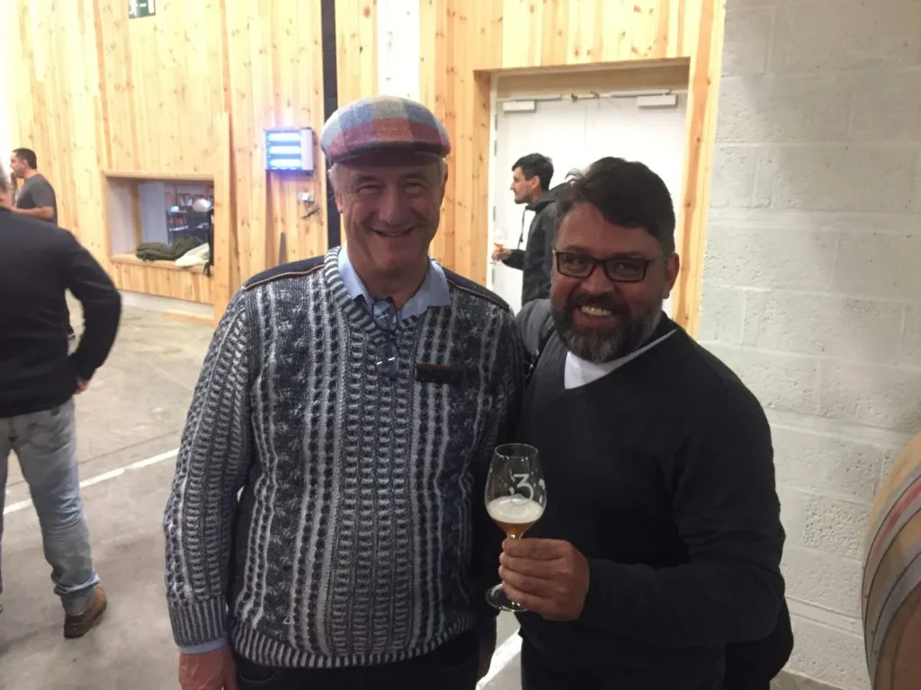 Visitante posa com Armand Debelder na 3 Fonteinen, segurando taça de cerveja com a marca 3
