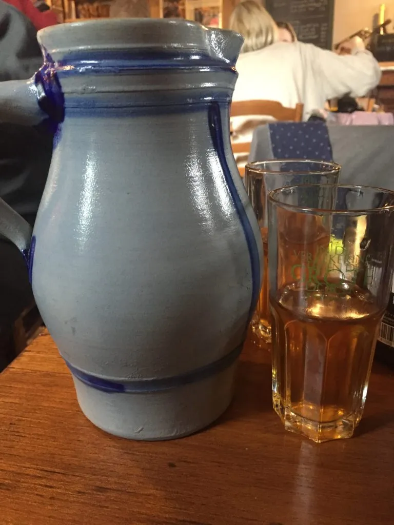 Jarra de cerâmica cinza com detalhes azuis ao lado de copos com Lambic puro, sobre mesa de madeira